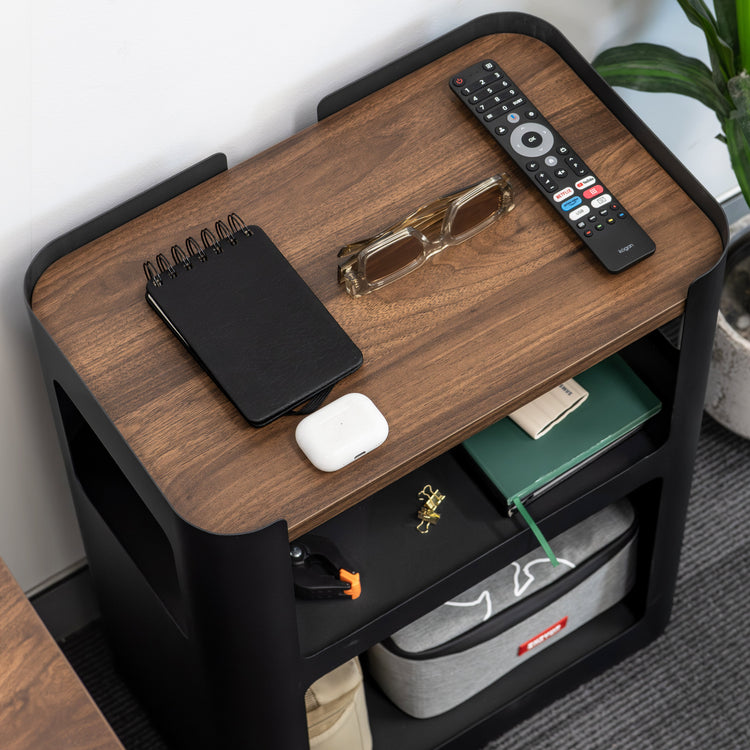 Wooden side table with remote control, phone, and other items on a gray carpeted floor.